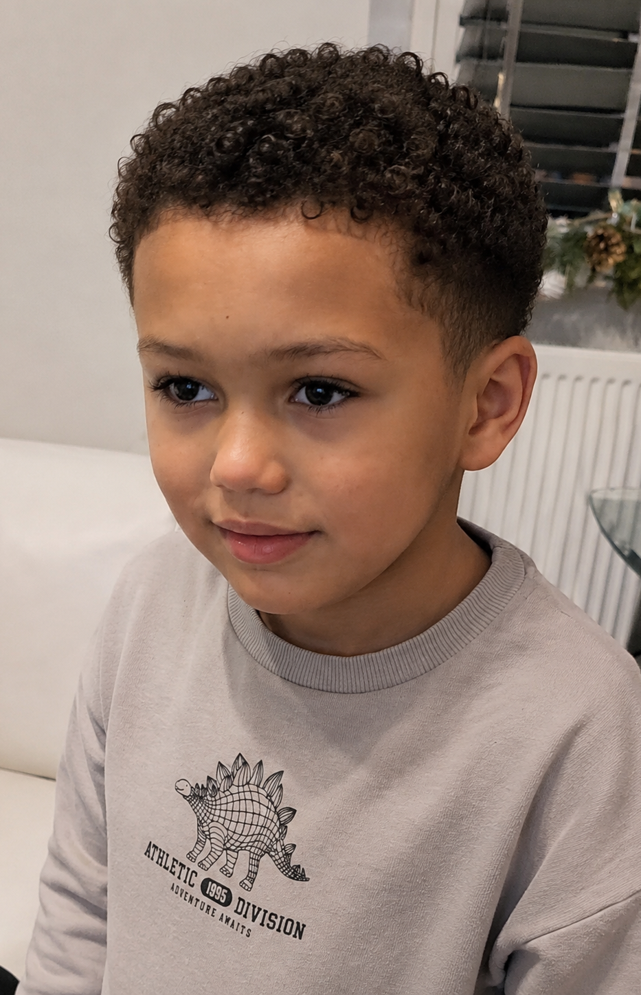 haircuts for mixed race boys hair with ASD, autism in London A young boy with curly hair, wearing a grey sweater, looking thoughtfully off to the side.